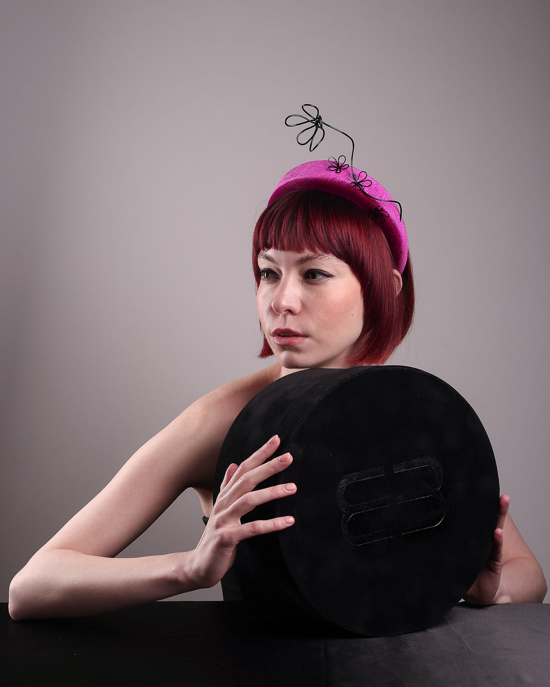Editorial picture of the model holding to a Merve Bayindir hat box while showcasing the front right side of a magenta base, features the Arcana high-formed headband, which sits neatly against the head. At the same time, the matte black floral wire detail ascends upward, accompanied by black wire flowers that create a doodle-like illusion. Designed by Merve Bayindir