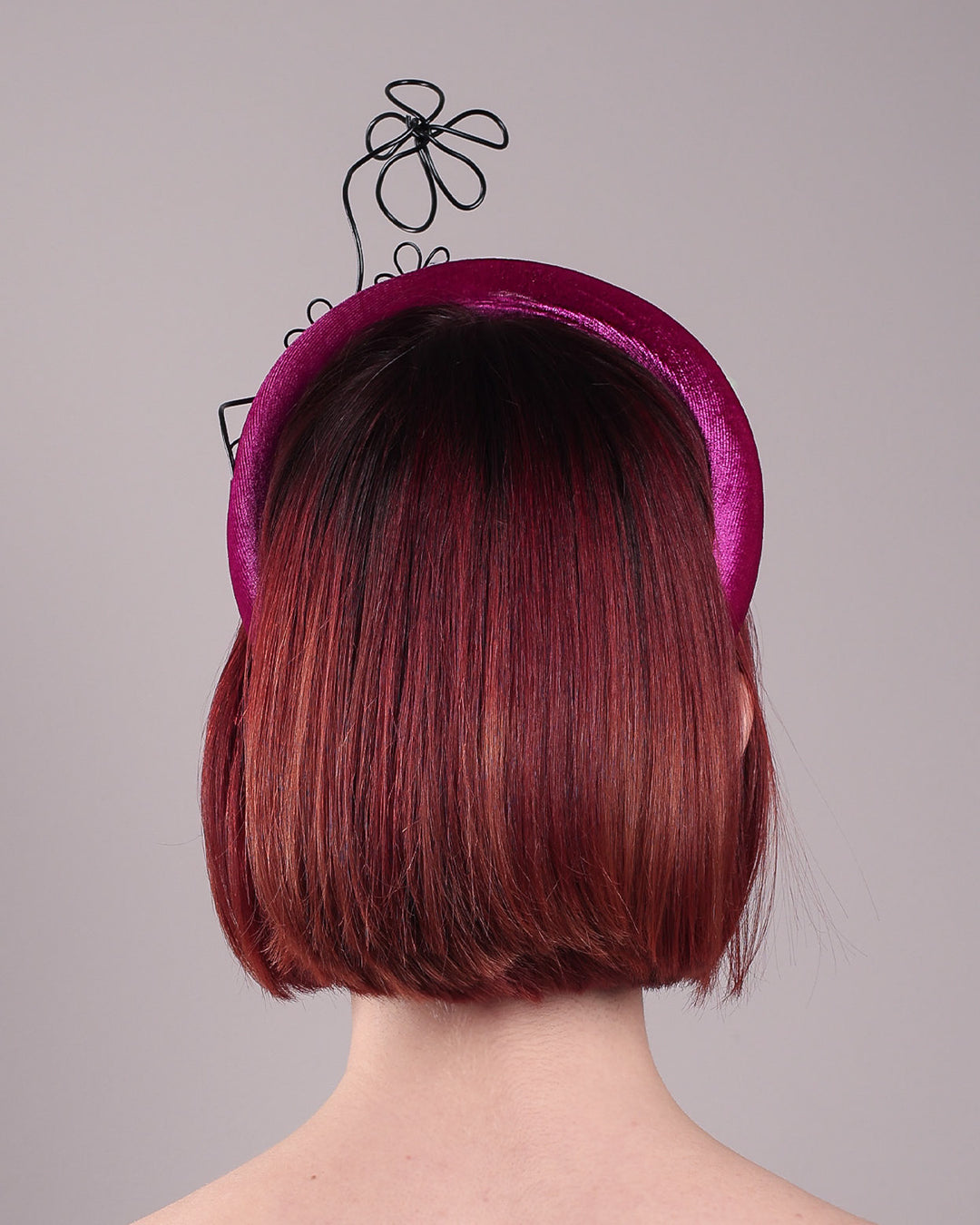 The model, showcasing the back of a magenta base, features the Arcana high-formed headband, which sits neatly against the head. At the same time, the matte black floral wire detail ascends upward, accompanied by black wire flowers that create a doodle-like illusion. Designed by Merve Bayindir