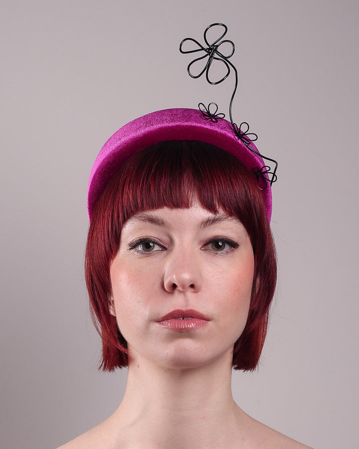 The model, showcasing the front close up of a magenta base, features the Arcana high-formed headband, which sits neatly against the head. At the same time, the matte black floral wire detail ascends upward, accompanied by black wire flowers that create a doodle-like illusion. Designed by Merve Bayindir