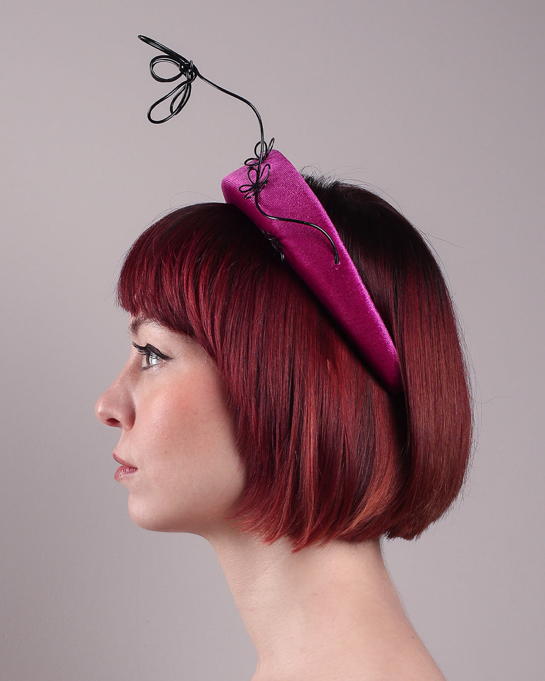 The model, showcasing the left side close up of a magenta base, features the Arcana high-formed headband, which sits neatly against the head. At the same time, the matte black floral wire detail ascends upward, accompanied by black wire flowers that create a doodle-like illusion. Designed by Merve Bayindir