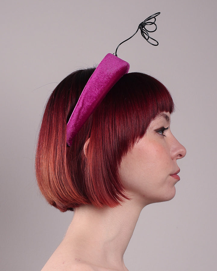 The model, showcasing the right side of a magenta base, features the Arcana high-formed headband, which sits neatly against the head. At the same time, the matte black floral wire detail ascends upward, accompanied by black wire flowers that create a doodle-like illusion. Designed by Merve Bayindir