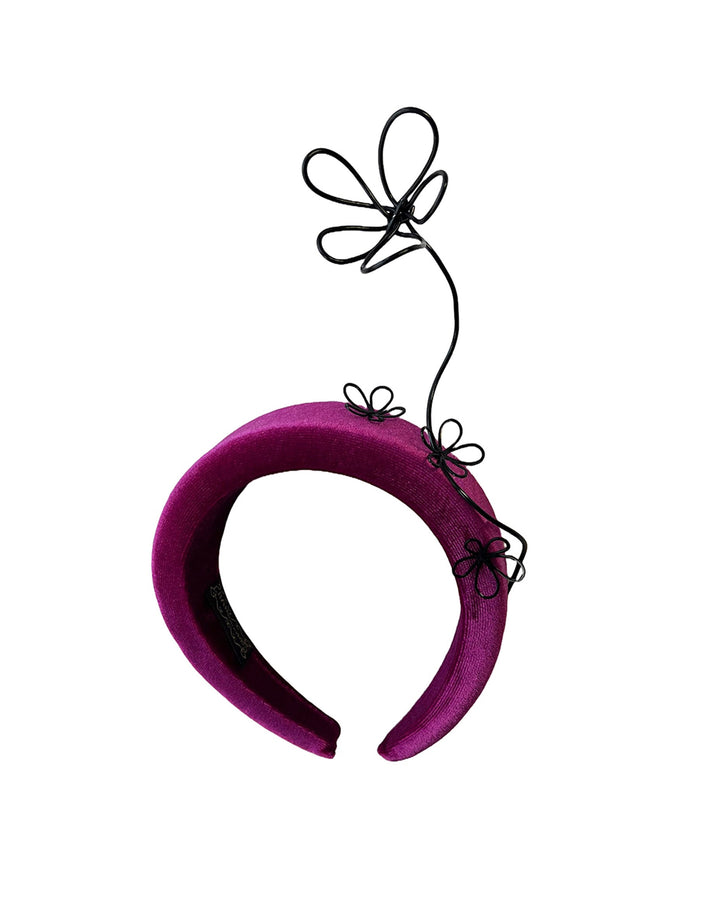 Still life picture of a magenta base, features the Arcana high-formed headband, which sits neatly against the head. At the same time, the matte black floral wire detail ascends upward, accompanied by black wire flowers that create a doodle-like illusion. Designed by Merve Bayindir