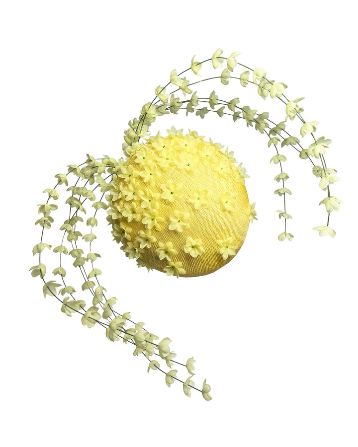 Bright yellow sinamay base fascinator shaped in a smooth rounded form, adorned with clusters of small soft-yellow flowers and framed by airy trailing floral stems that curve gracefully around the piece. Designed and made by Merve Bayindir Millinery.

