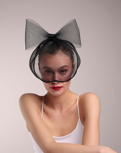 The model is wearing a black mesh mask covering almost over face, leaving the lips and chin visible. On top of the cage-style mesh mask, there is a mesh bow. The mask is perfect for high fashion events or special parties.