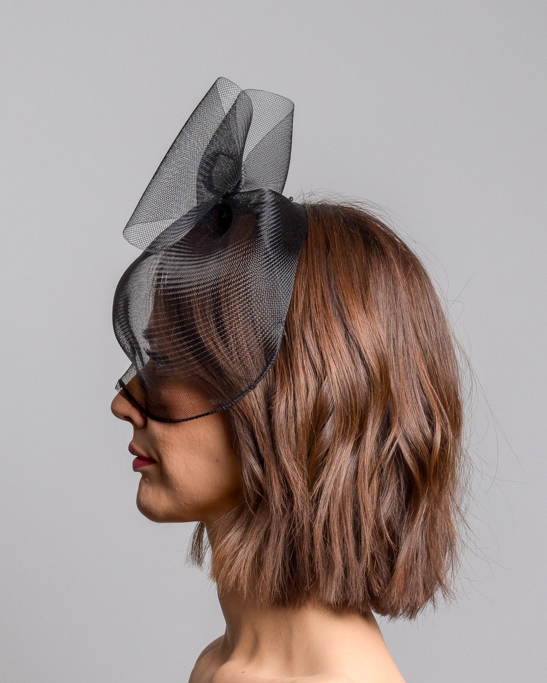 Model is showcasing the left side of the black mesh cage-style constructed Astrea mask. On top of the cage-style mesh mask, there is a mesh bow. The mask is fitted with a thin headband. The mask is perfect for high-fashion events or special parties. Made by Merve Bayindir Millinery.