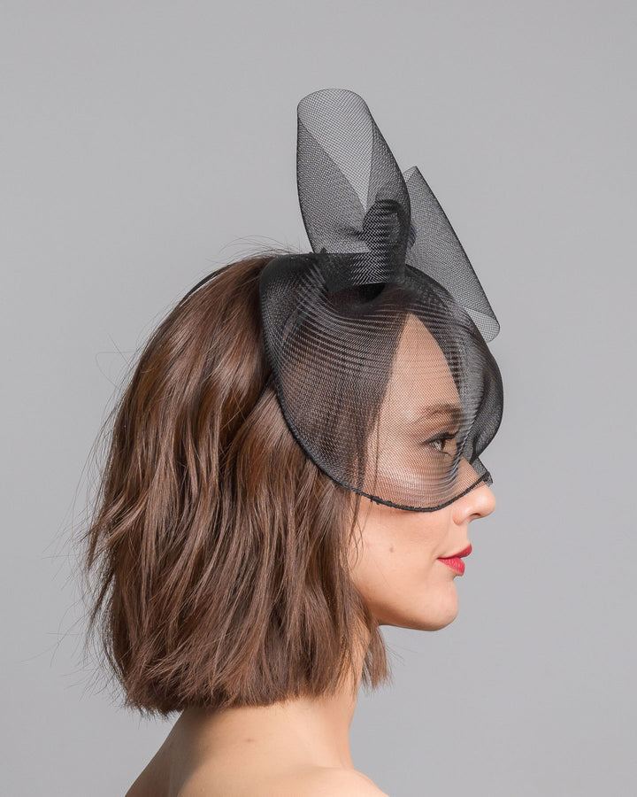 Model is showcasing the right side of the black mesh cage-style constructed Astrea mask. On top of the cage-style mesh mask, there is a mesh bow. The mask is fitted with a thin headband. The mask is perfect for high-fashion events or special parties. Made by Merve Bayindir Millinery.