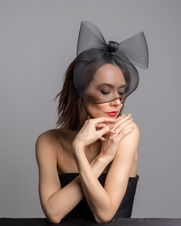 Model is posing wearing the black mesh cage-style constructed Astrea mask. On top of the cage-style mesh mask, there is a mesh bow. The mask is fitted with a thin headband. The mask is perfect for high-fashion events or special parties. Made by Merve Bayindir Millinery.