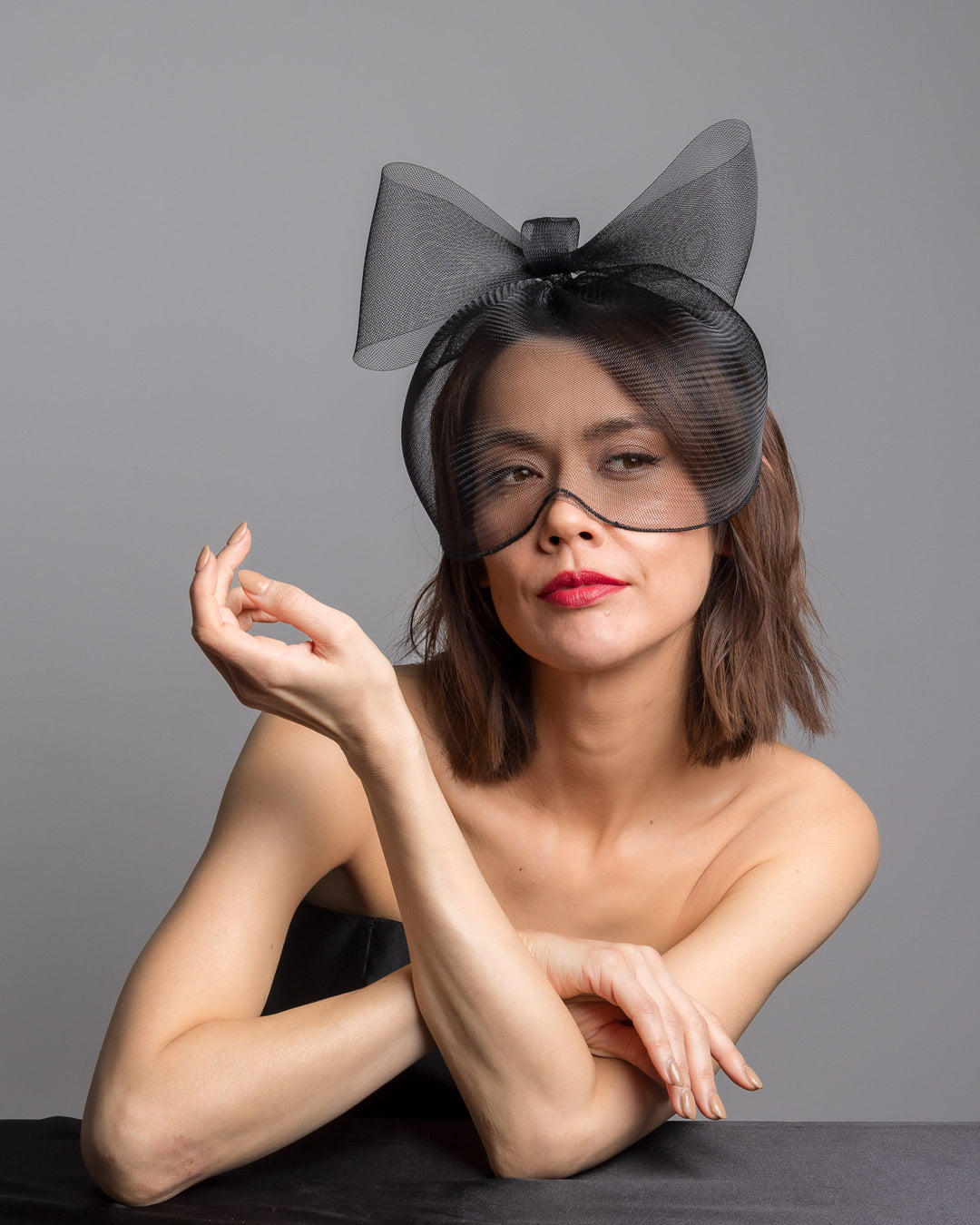 Model is posing wearing the black mesh cage-style constructed Astrea mask. On top of the cage-style mesh mask, there is a mesh bow. The mask is fitted with a thin headband. The mask is perfect for high-fashion events or special parties. Made by Merve Bayindir Millinery.
