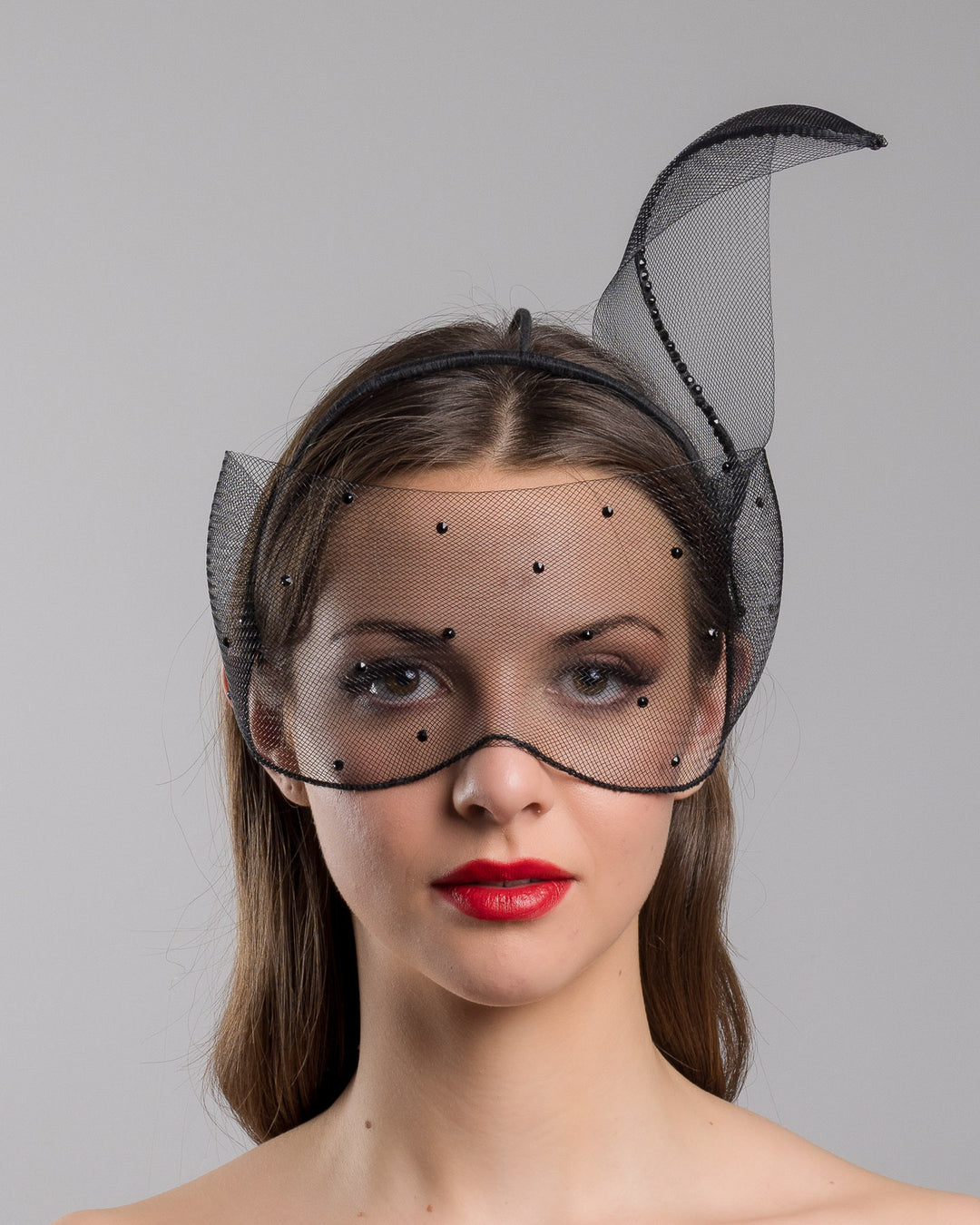 Model is showcasing the front of the black mesh headpiece is adorned with small black crystals. It features a design with a prominent bow-like structure on one side embellished with black crystals. The delicate mesh has a wire band to be fitted to the head. Made by Merve Bayindir Millinery.