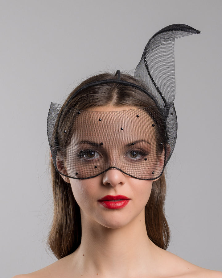 Model is showcasing the front of the black mesh headpiece is adorned with small black crystals. It features a design with a prominent bow-like structure on one side embellished with black crystals. The delicate mesh has a wire band to be fitted to the head. Made by Merve Bayindir Millinery.