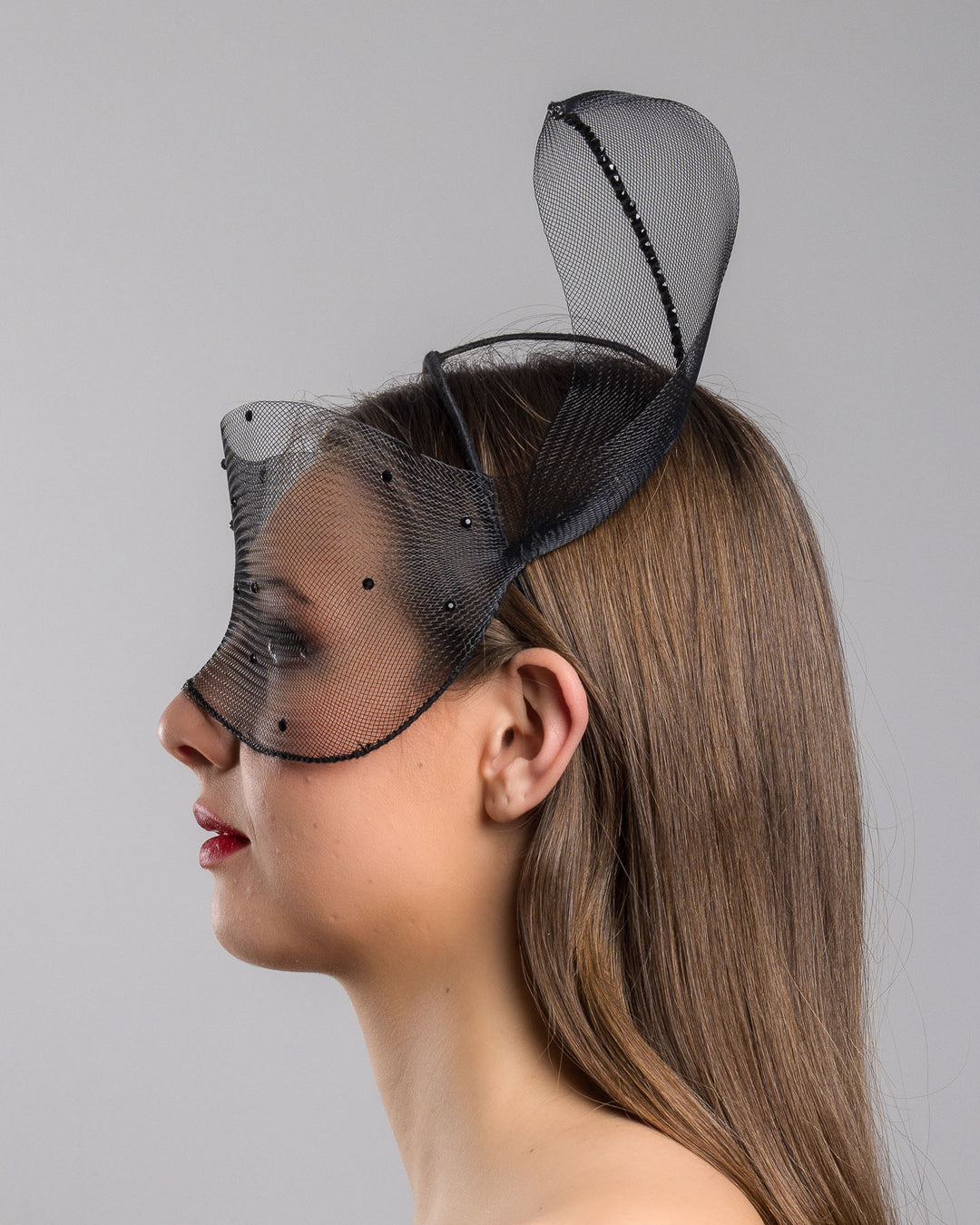 Model is showcasing the left side of the black mesh headpiece is adorned with small black crystals. It features a design with a prominent bow-like structure on one side embellished with black crystals. The delicate mesh has a wire band to be fitted to the head. Made by Merve Bayindir Millinery.