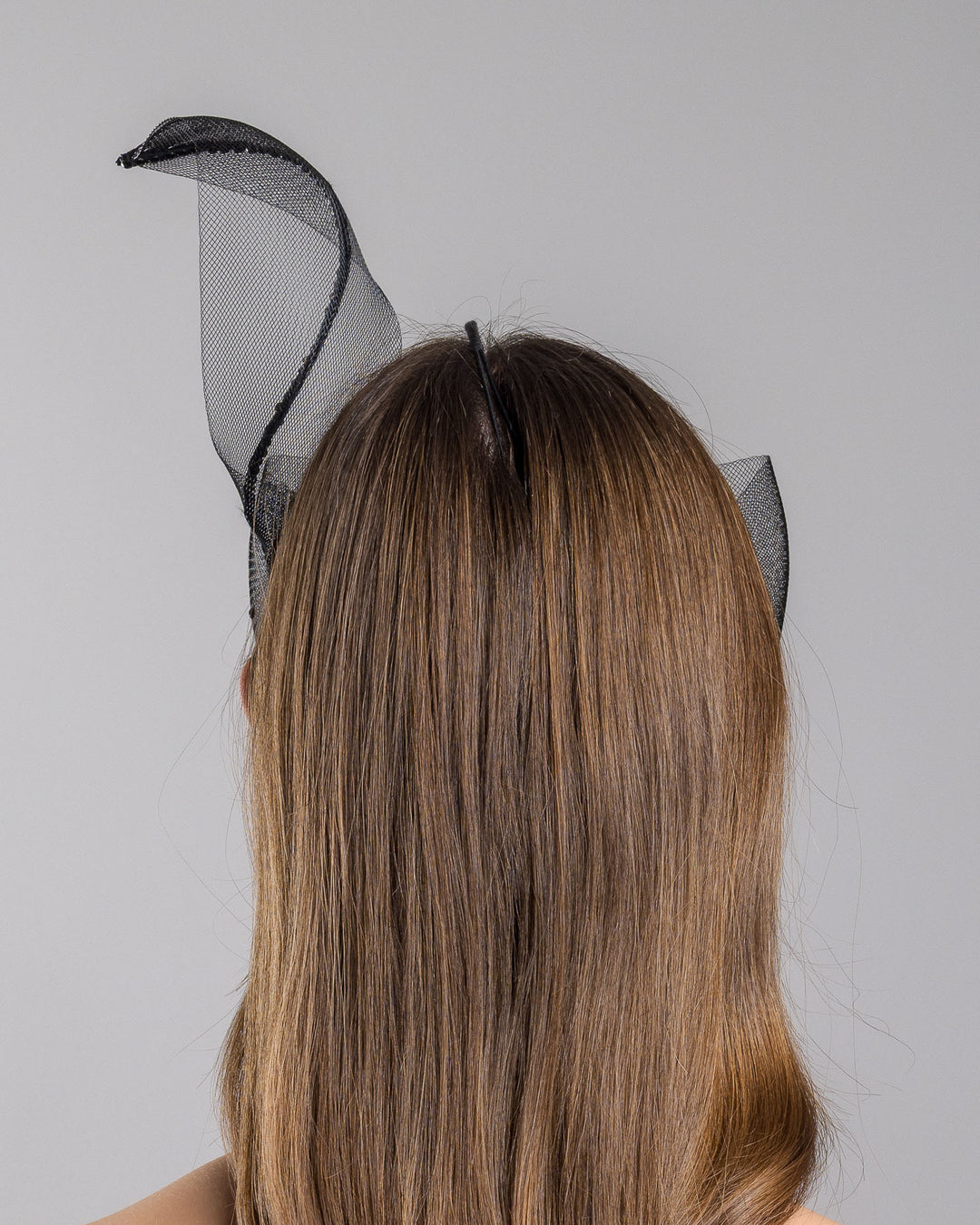 Model is showcasing the back of the black mesh headpiece is adorned with small black crystals. It features a design with a prominent bow-like structure on one side embellished with black crystals. The delicate mesh has a wire band to be fitted to the head. Made by Merve Bayindir Millinery.