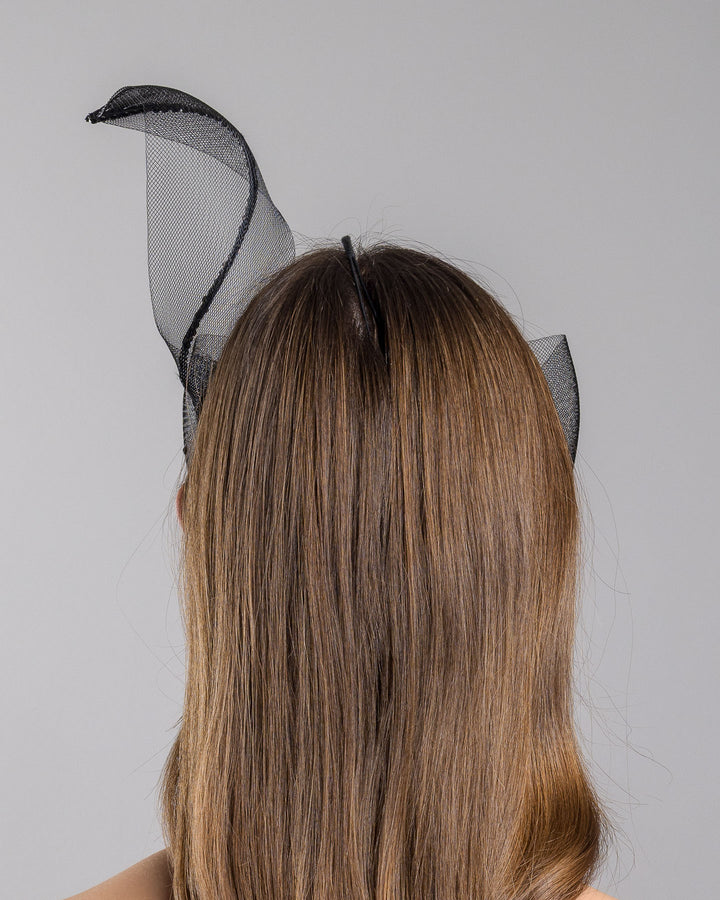 Model is showcasing the back of the black mesh headpiece is adorned with small black crystals. It features a design with a prominent bow-like structure on one side embellished with black crystals. The delicate mesh has a wire band to be fitted to the head. Made by Merve Bayindir Millinery.