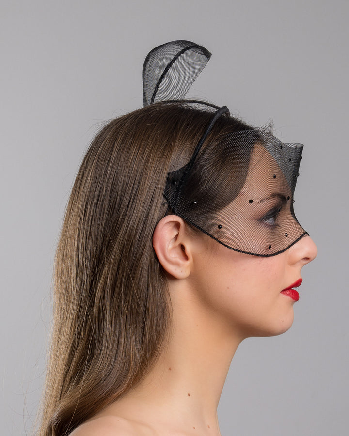Model is showcasing the right side of the black mesh headpiece is adorned with small black crystals. It features a design with a prominent bow-like structure on one side embellished with black crystals. The delicate mesh has a wire band to be fitted to the head. Made by Merve Bayindir Millinery.