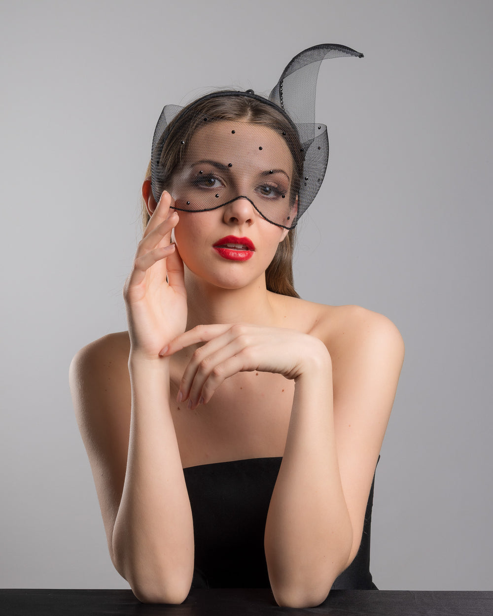 Model is posing wearing the Atlanta mask, the black mesh headpiece is adorned with small black crystals. It features a design with a prominent bow-like structure on one side embellished with black crystals. The delicate mesh has a wire band to be fitted to the head. Made by Merve Bayindir Millinery.