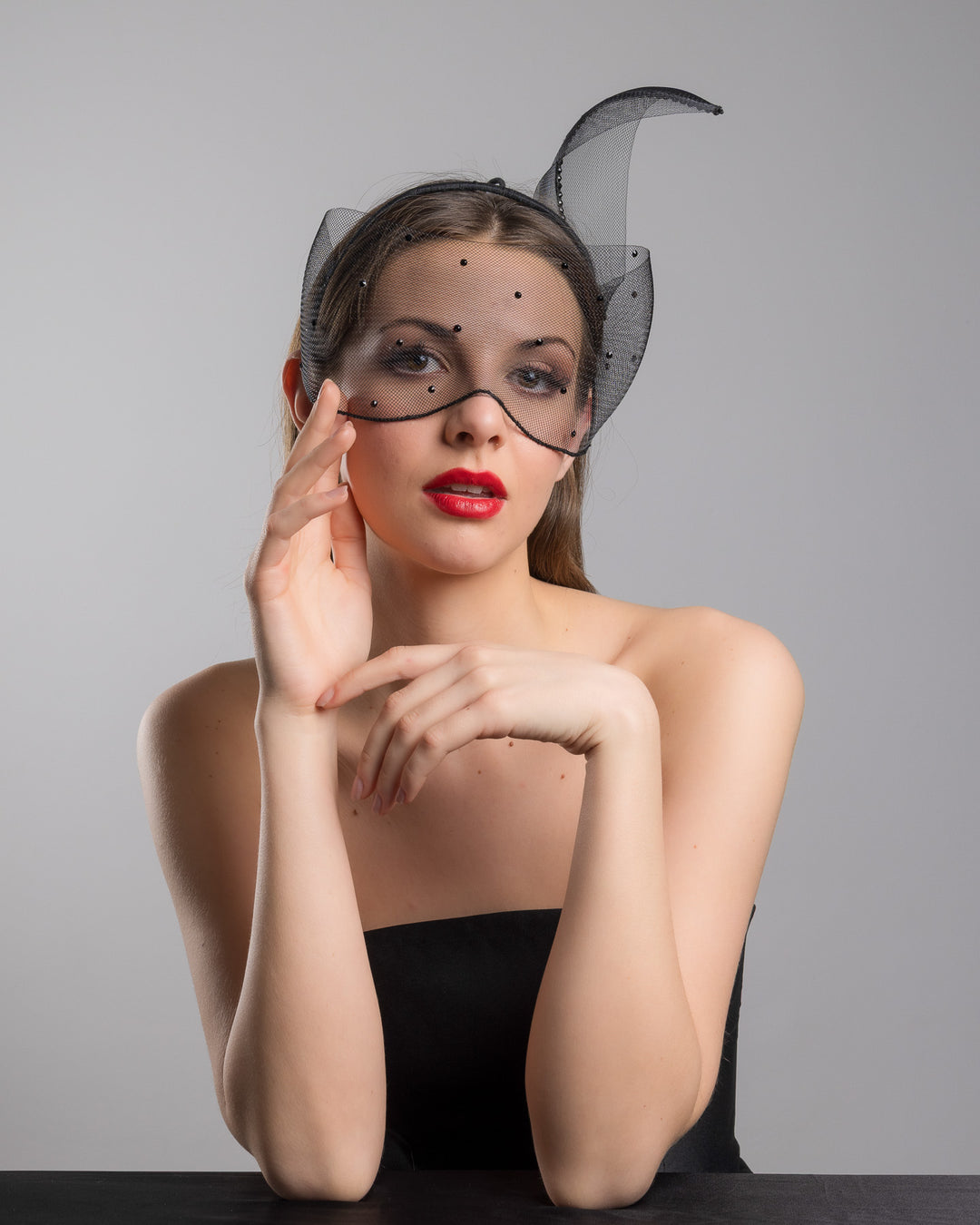 Model is posing wearing the Atlanta mask, the black mesh headpiece is adorned with small black crystals. It features a design with a prominent bow-like structure on one side embellished with black crystals. The delicate mesh has a wire band to be fitted to the head. Made by Merve Bayindir Millinery.