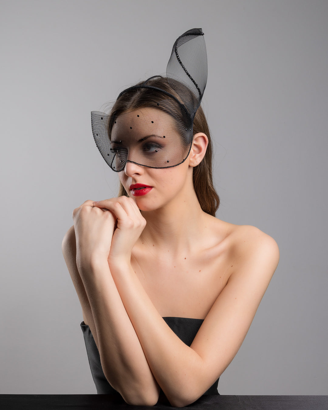 Model is posing wearing the Atlanta mask, the black mesh headpiece is adorned with small black crystals. It features a design with a prominent bow-like structure on one side embellished with black crystals. The delicate mesh has a wire band to be fitted to the head. Made by Merve Bayindir Millinery.