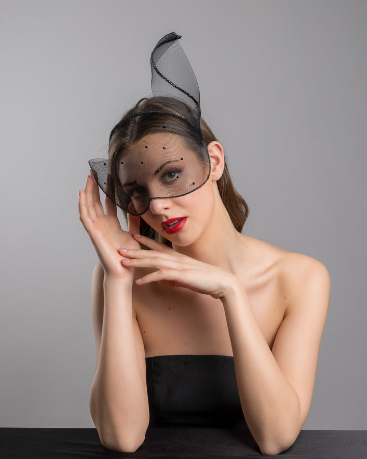 Model is posing wearing the Atlanta mask, the black mesh headpiece is adorned with small black crystals. It features a design with a prominent bow-like structure on one side embellished with black crystals. The delicate mesh has a wire band to be fitted to the head. Made by Merve Bayindir Millinery.
