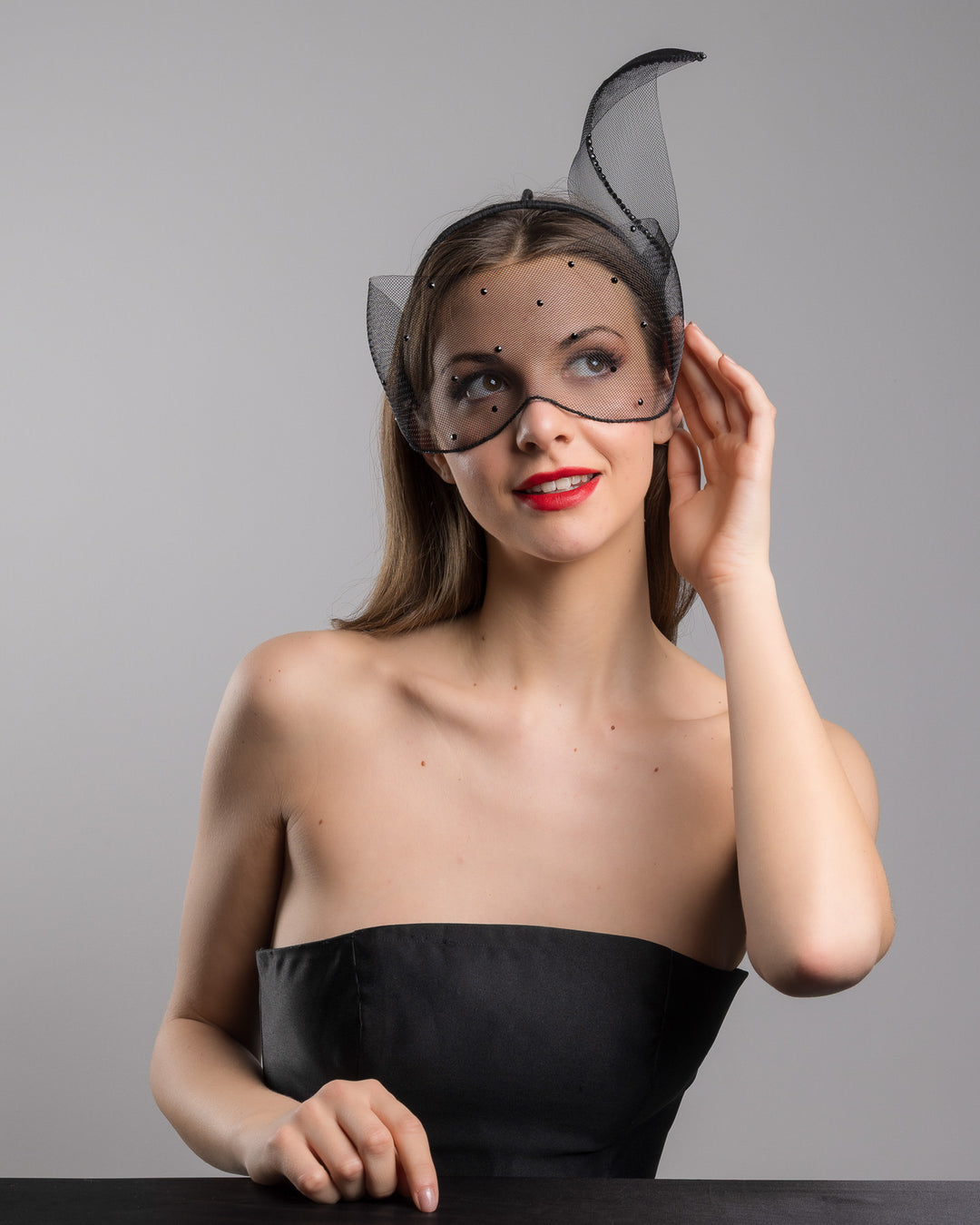 Model is posing wearing the Atlanta mask, the black mesh headpiece is adorned with small black crystals. It features a design with a prominent bow-like structure on one side embellished with black crystals. The delicate mesh has a wire band to be fitted to the head. Made by Merve Bayindir Millinery.