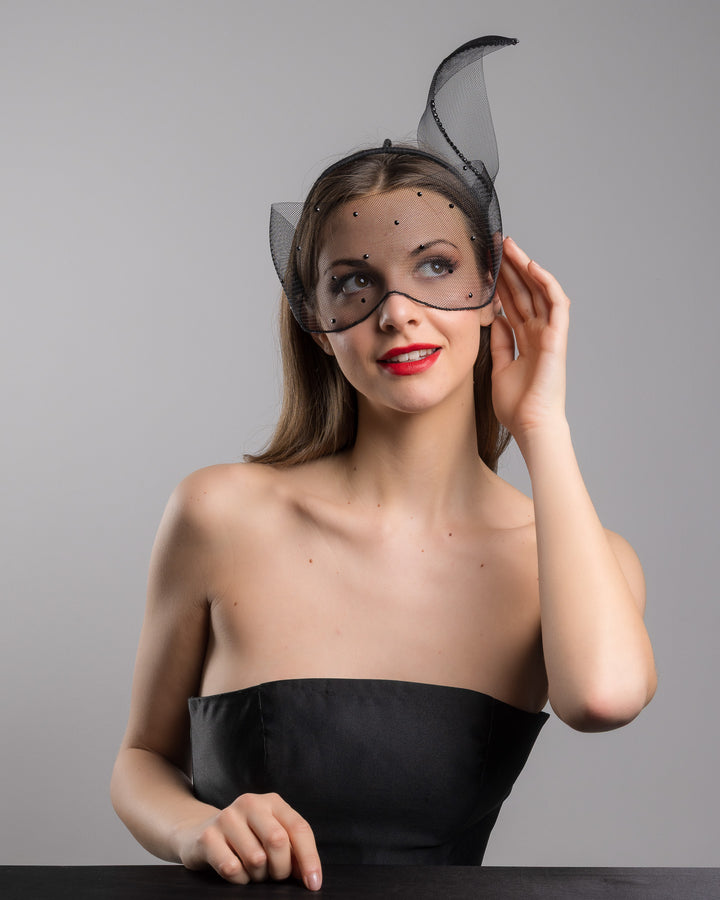 Model is posing wearing the Atlanta mask, the black mesh headpiece is adorned with small black crystals. It features a design with a prominent bow-like structure on one side embellished with black crystals. The delicate mesh has a wire band to be fitted to the head. Made by Merve Bayindir Millinery.