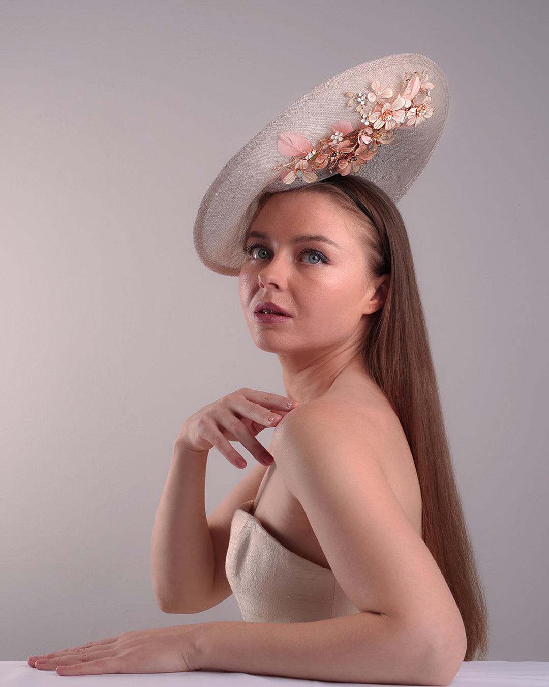 Model is showcasing the right below angle of a saucer style grey hat fitted on a small saucer piece to give a taller reach worn tilted to the right side of the head. The left high side has curve creating a large space on the left inside. The inside part of the hat is decorated with resin and crystal mixed flower arrangement, embellished with trimmed feathers. Fitted with a wire headband and elastic.