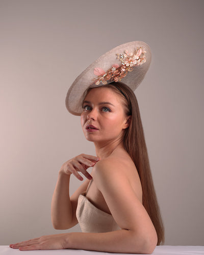 Model is showcasing the right below angle of a saucer style grey hat fitted on a small saucer piece to give a taller reach worn tilted to the right side of the head. The left high side has curve creating a large space on the left inside. The inside part of the hat is decorated with resin and crystal mixed flower arrangement, embellished with trimmed feathers. Fitted with a wire headband and elastic.