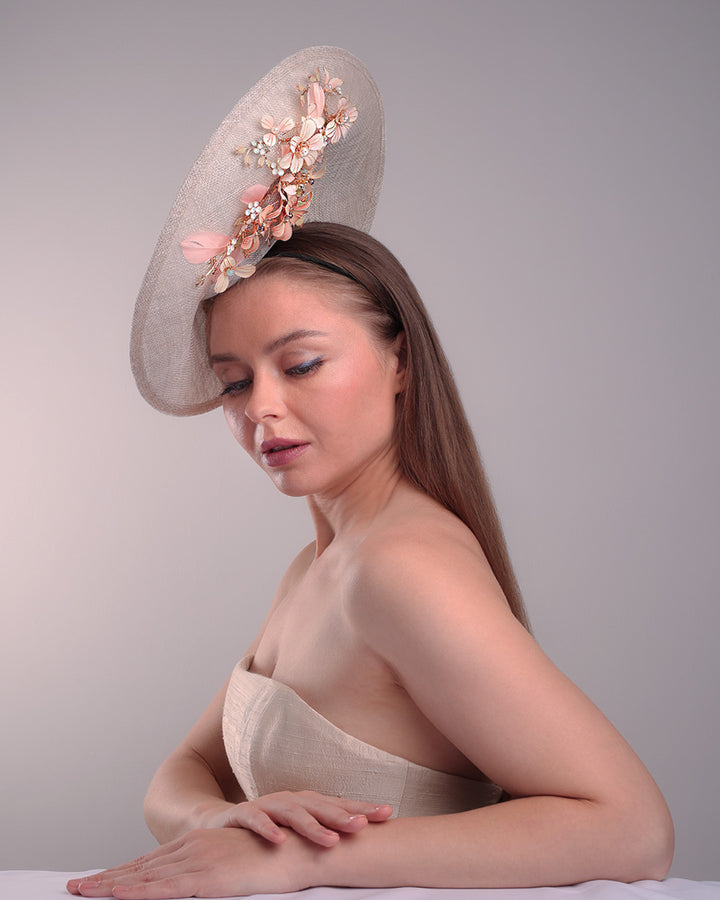 Model is showcasing the right high angle of a saucer style grey hat fitted on a small saucer piece to give a taller reach worn tilted to the right side of the head. The left high side has curve creating a large space on the left inside. The inside part of the hat is decorated with resin and crystal mixed flower arrangement, embellished with trimmed feathers. Fitted with a wire headband and elastic.
