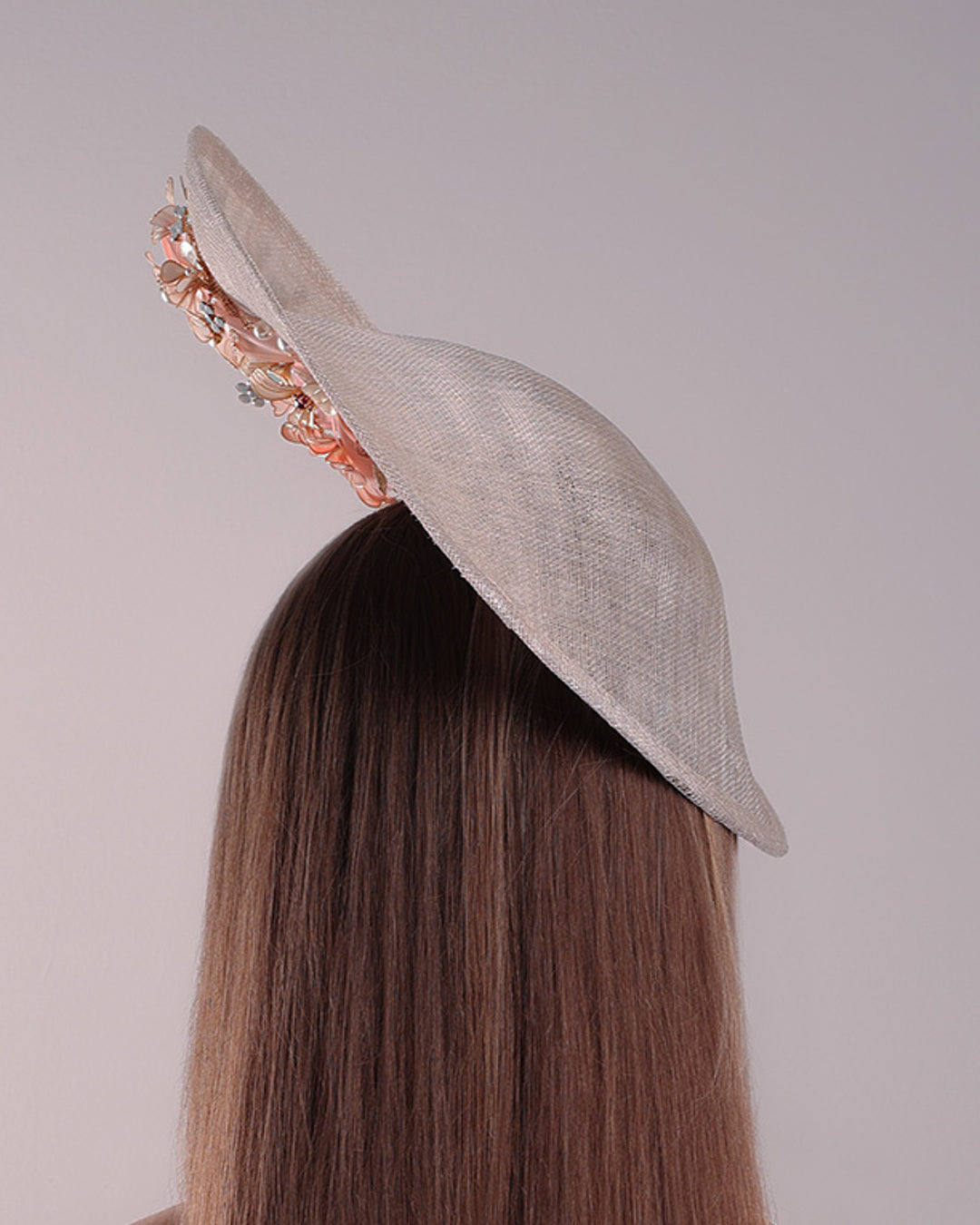 Model is showcasing the back of a saucer style grey hat fitted on a small saucer piece to give a taller reach worn tilted to the right side of the head. The left high side has curve creating a large space on the left inside. The inside part of the hat is decorated with resin and crystal mixed flower arrangement, embellished with trimmed feathers. Fitted with a wire headband and elastic.