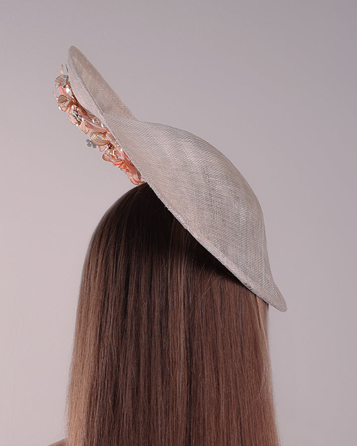 Model is showcasing the back of a saucer style grey hat fitted on a small saucer piece to give a taller reach worn tilted to the right side of the head. The left high side has curve creating a large space on the left inside. The inside part of the hat is decorated with resin and crystal mixed flower arrangement, embellished with trimmed feathers. Fitted with a wire headband and elastic.