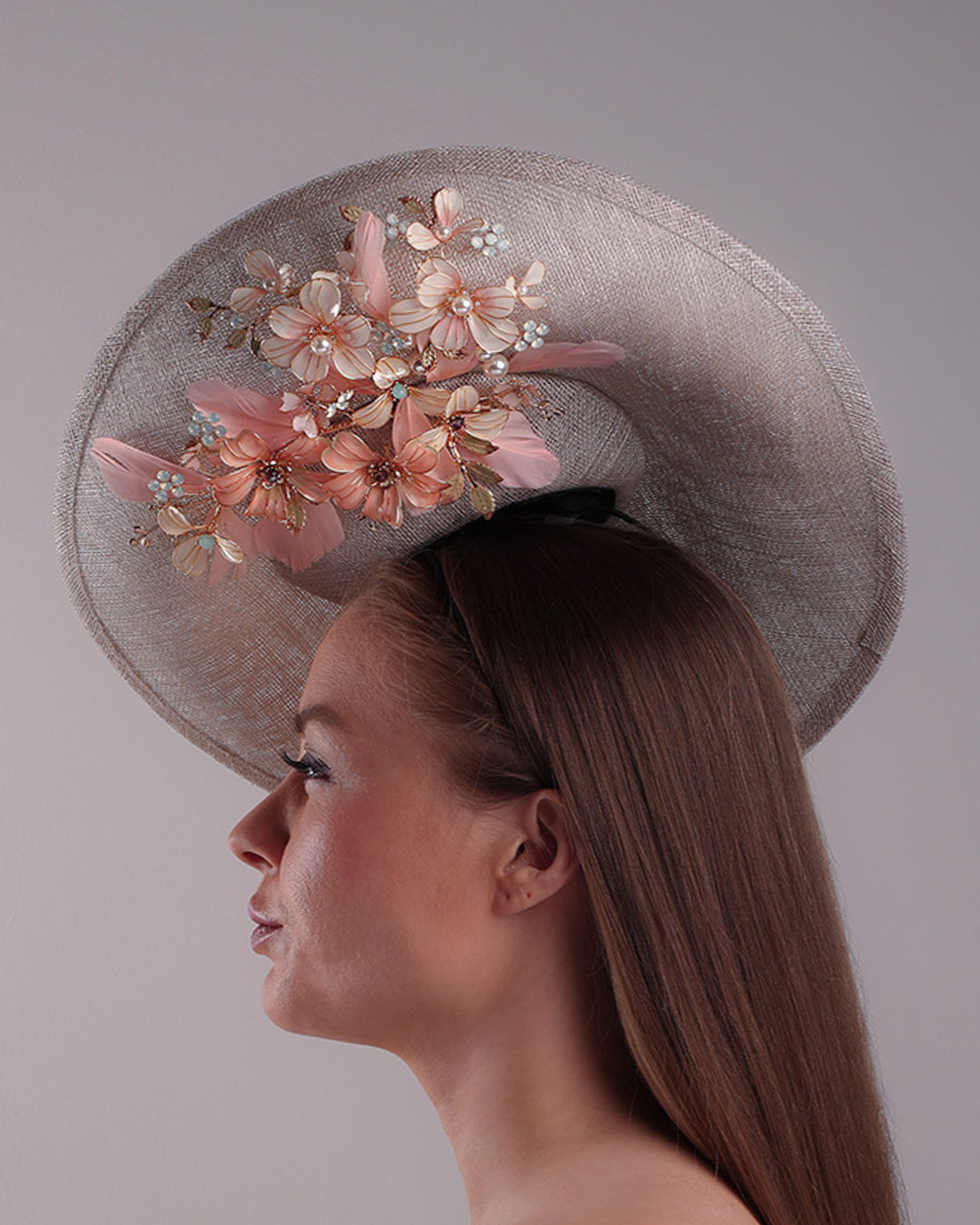 Model is showcasing the left side of a saucer style grey hat fitted on a small saucer piece to give a taller reach worn tilted to the right side of the head. The left high side has curve creating a large space on the left inside. The inside part of the hat is decorated with resin and crystal mixed flower arrangement, embellished with trimmed feathers. Fitted with a wire headband and elastic.