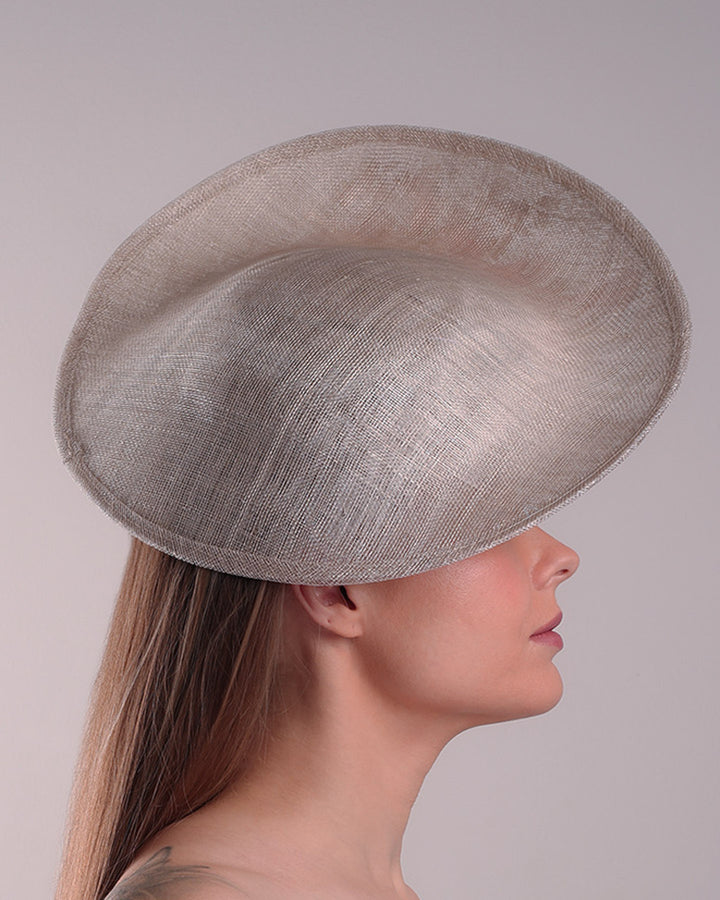 Model is showcasing the right side of a saucer style grey hat fitted on a small saucer piece to give a taller reach worn tilted to the right side of the head. The left high side has curve creating a large space on the left inside. The inside part of the hat is decorated with resin and crystal mixed flower arrangement, embellished with trimmed feathers. Fitted with a wire headband and elastic.