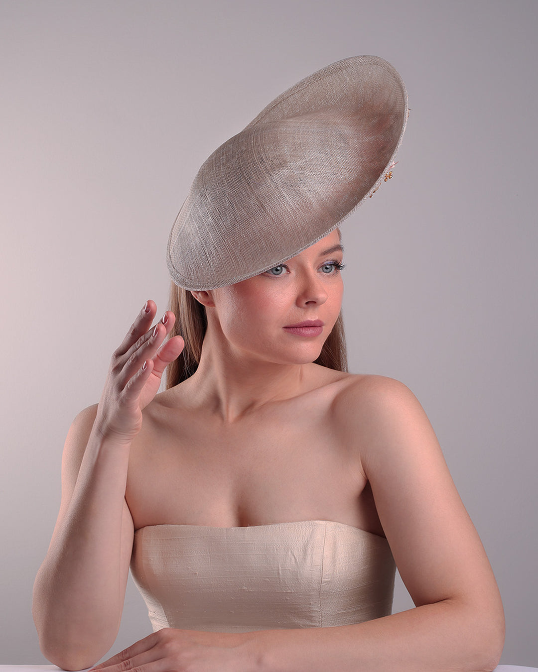 Model is showcasing the left front angle of a saucer style grey hat fitted on a small saucer piece to give a taller reach worn tilted to the right side of the head. The left high side has curve creating a large space on the left inside. The inside part of the hat is decorated with resin and crystal mixed flower arrangement, embellished with trimmed feathers. Fitted with a wire headband and elastic.