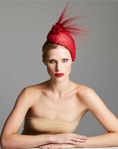 The model wears a fascinator, a vibrant red headpiece designed to sit elegantly on the side of the head. It features a quilted texture over a sinamay and is adorned with a cluster of delicate red roses at the top. A dramatic spray of red feathers extends upward and outward, adding height and flair to the accessory. The model wearing it has a sleek, pulled-back hairstyle and striking red lipstick, complementing the fascinator’s bold colour..