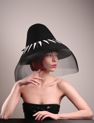 The model showcases the front right side of the Ava hat, featuring an exaggerated, proportioned sculptural high-coned silhouette crafted in jet-black felt and crowned with sharp white and black feather accents. A sheer black crinoline veil spills from the brim. Designed by Merve Bayindir.