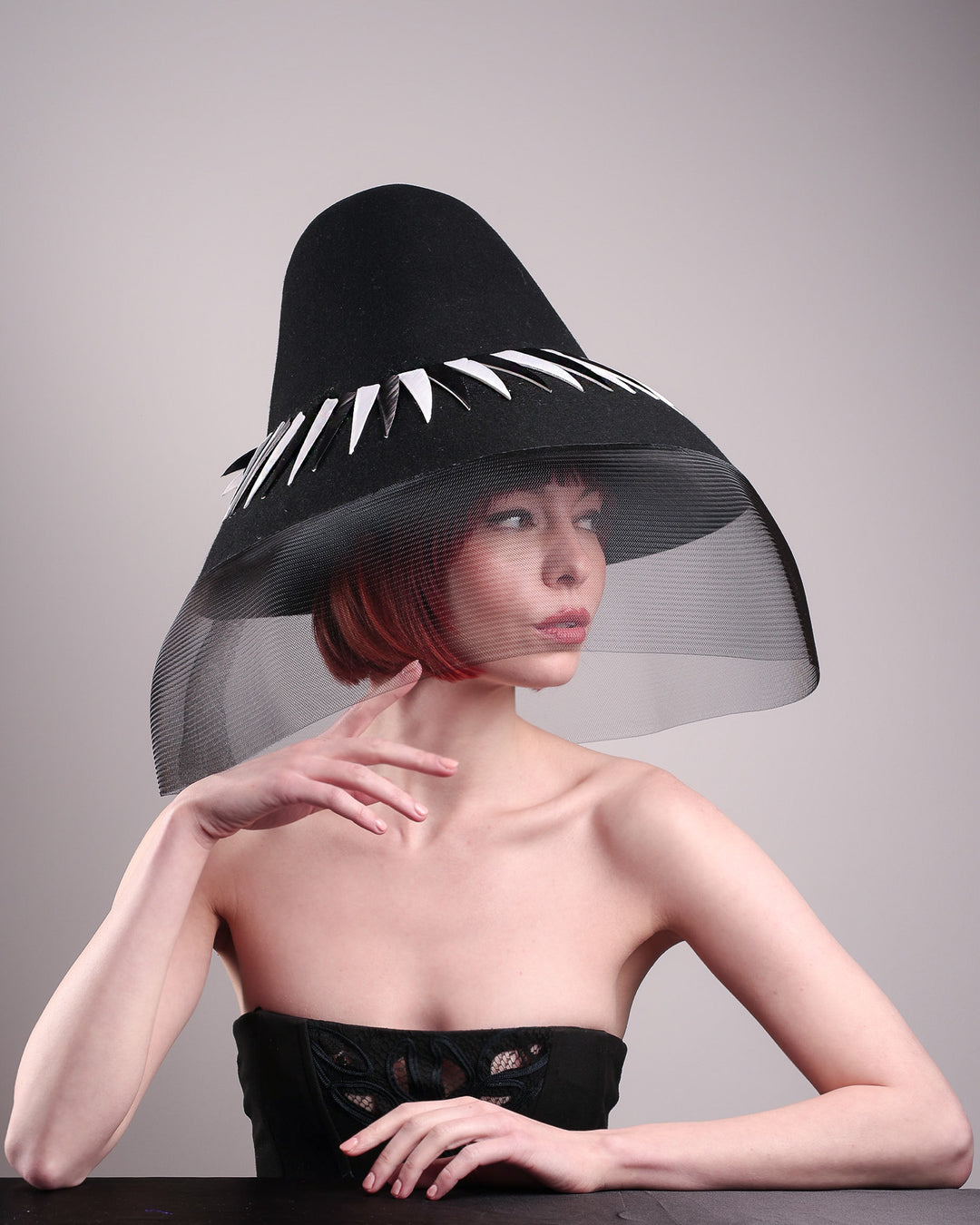 The model showcases the front right side of the Ava hat, featuring an exaggerated, proportioned sculptural high-coned silhouette crafted in jet-black felt and crowned with sharp white and black feather accents. A sheer black crinoline veil spills from the brim. Designed by Merve Bayindir.