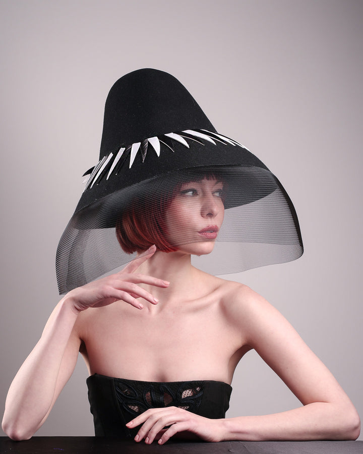 The model showcases the front right side of the Ava hat, featuring an exaggerated, proportioned sculptural high-coned silhouette crafted in jet-black felt and crowned with sharp white and black feather accents. A sheer black crinoline veil spills from the brim. Designed by Merve Bayindir.