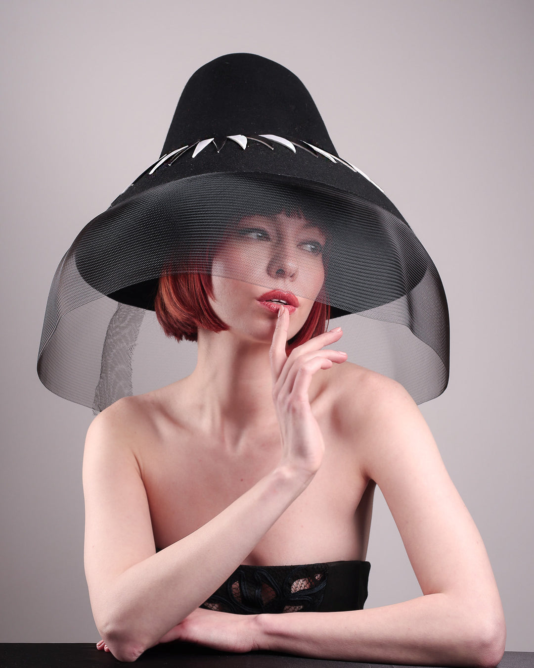 The model showcases the lower angle front view of the Ava hat, featuring an exaggerated, proportioned sculptural high-coned silhouette crafted in jet-black felt and crowned with sharp white and black feather accents. A sheer black crinoline veil spills from the brim. Designed by Merve Bayindir.