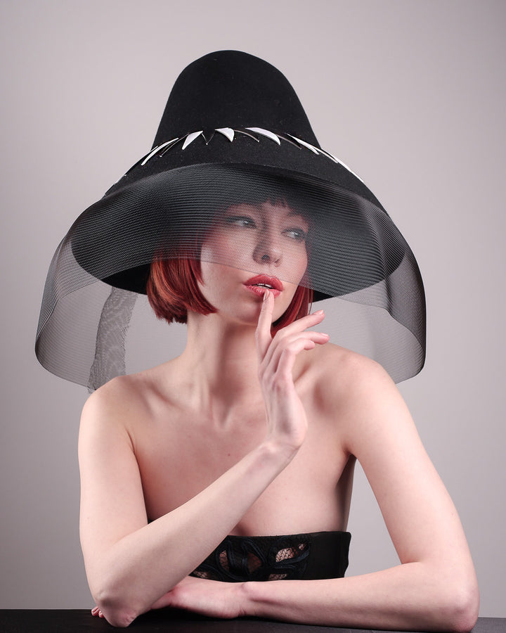 The model showcases the lower angle front view of the Ava hat, featuring an exaggerated, proportioned sculptural high-coned silhouette crafted in jet-black felt and crowned with sharp white and black feather accents. A sheer black crinoline veil spills from the brim. Designed by Merve Bayindir.