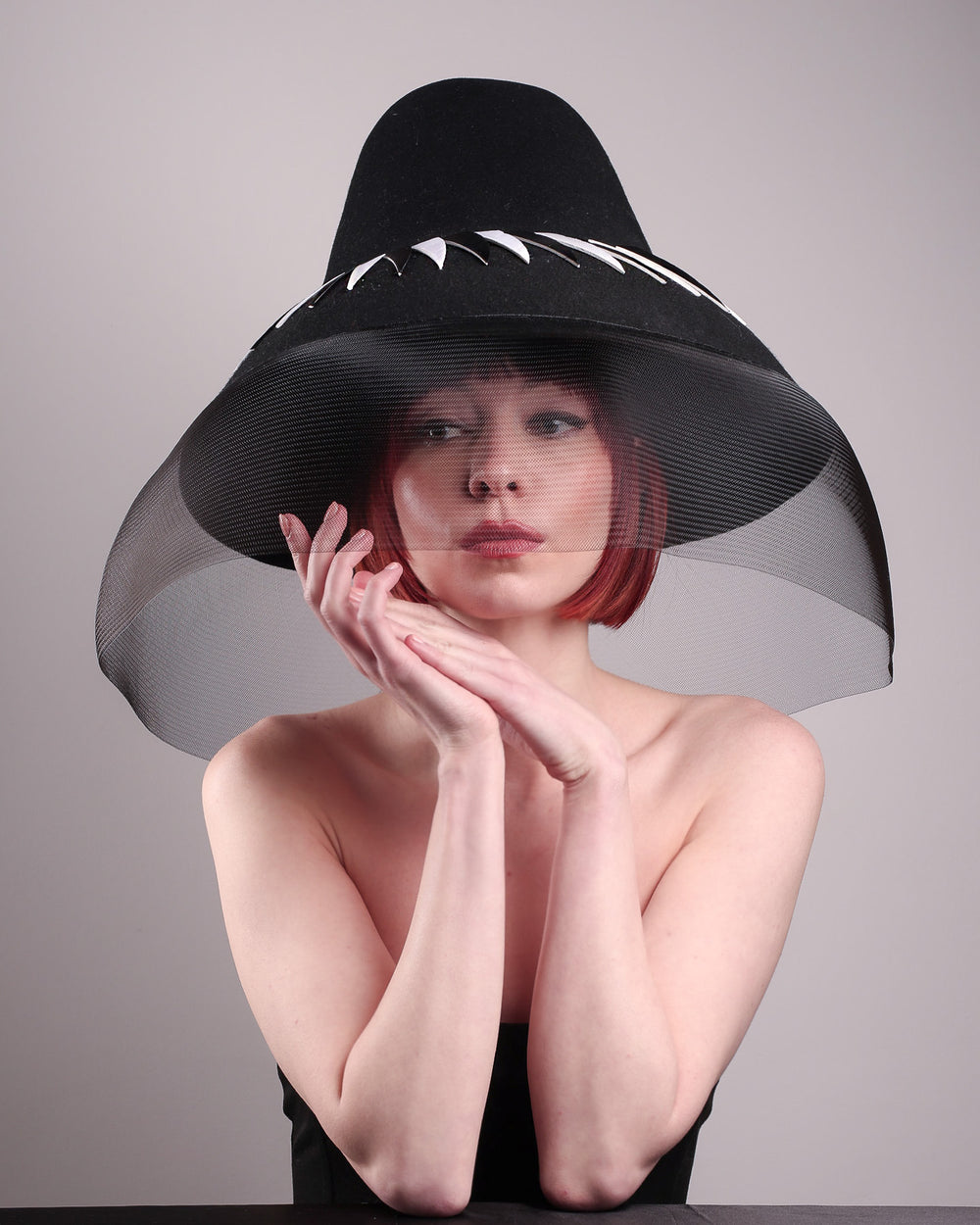 The model showcases the front of the Ava hat, featuring an exaggerated, proportioned sculptural high-coned silhouette crafted in jet-black felt and crowned with sharp white and black feather accents. A sheer black crinoline veil spills from the brim. Designed by Merve Bayindir.