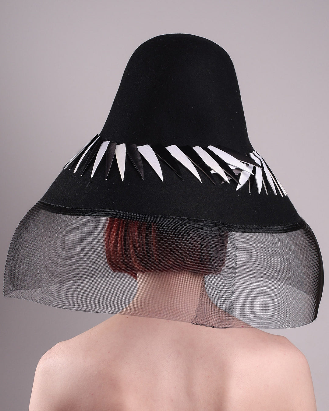 The model showcasing the back of the Ava hat, featuring an exaggerated, proportioned sculptural high-coned silhouette crafted in jet-black felt and crowned with sharp white and black feather accents. A sheer black crinoline veil spills from the brim. Designed by Merve Bayindir.