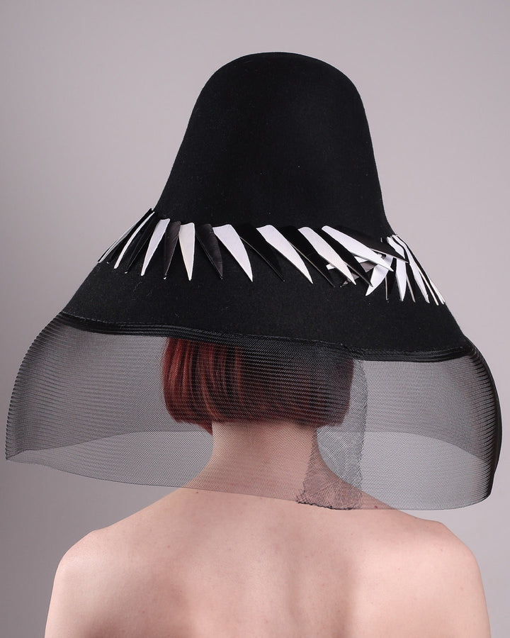 The model showcasing the back of the Ava hat, featuring an exaggerated, proportioned sculptural high-coned silhouette crafted in jet-black felt and crowned with sharp white and black feather accents. A sheer black crinoline veil spills from the brim. Designed by Merve Bayindir.