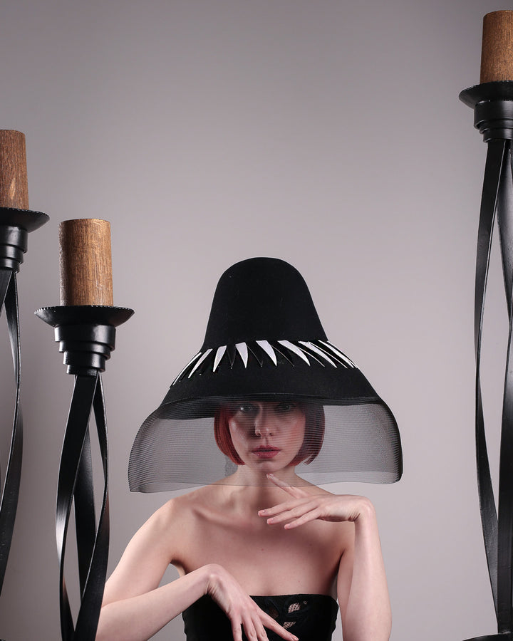 The model pooses between tall candles showcasing the front of the Ava hat, featuring an exaggerated, proportioned sculptural high-coned silhouette crafted in jet-black felt and crowned with sharp white and black feather accents. A sheer black crinoline veil spills from the brim. Designed by Merve Bayindir.