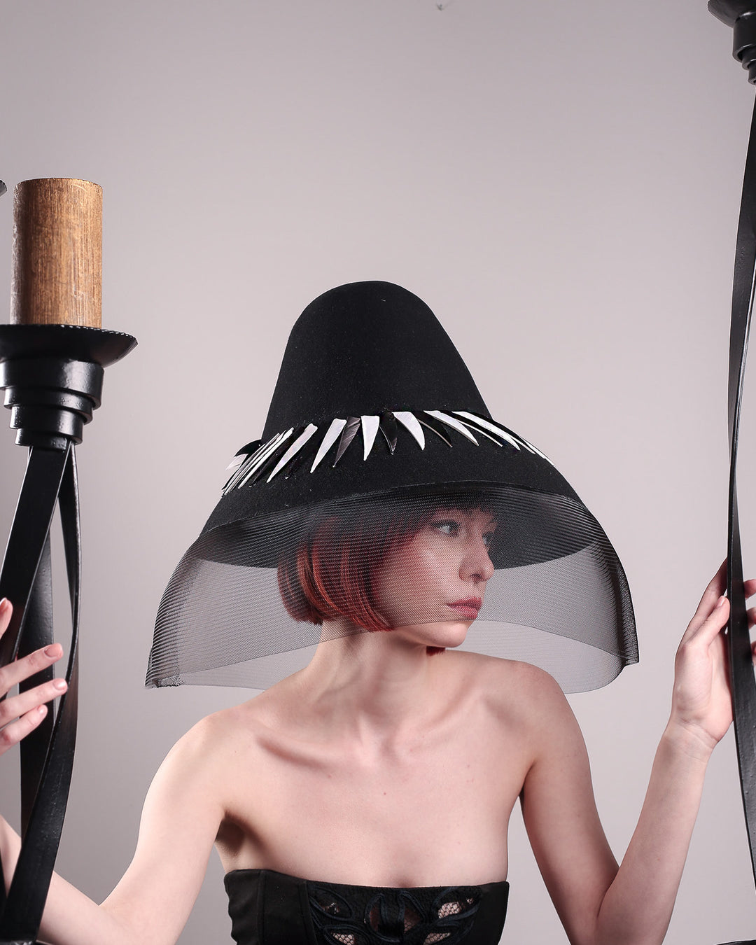 The model pooses between tall candles showcasing the front of the Ava hat, featuring an exaggerated, proportioned sculptural high-coned silhouette crafted in jet-black felt and crowned with sharp white and black feather accents. A sheer black crinoline veil spills from the brim. Designed by Merve Bayindir.
