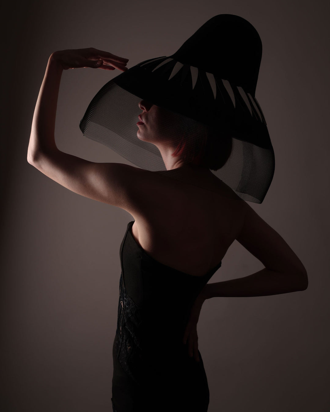 The model posing under the dark light showcasing theright side of the Ava hat, featuring an exaggerated, proportioned sculptural high-coned silhouette crafted in jet-black felt and crowned with sharp white and black feather accents. A sheer black crinoline veil spills from the brim. Designed by Merve Bayindir.