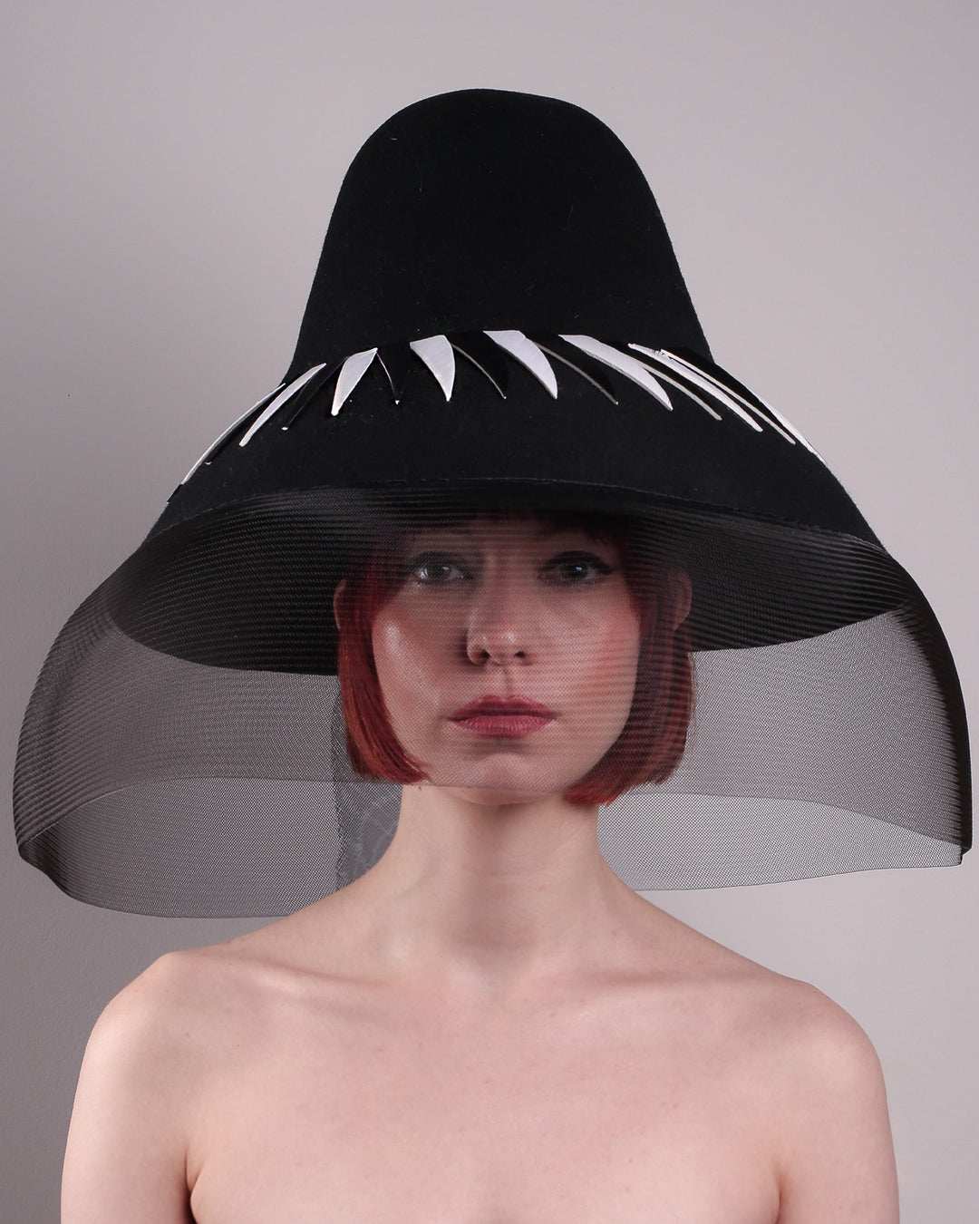 The model showcasing the front close up of the Ava hat, featuring an exaggerated, proportioned sculptural high-coned silhouette crafted in jet-black felt and crowned with sharp white and black feather accents. A sheer black crinoline veil spills from the brim. Designed by Merve Bayindir.
