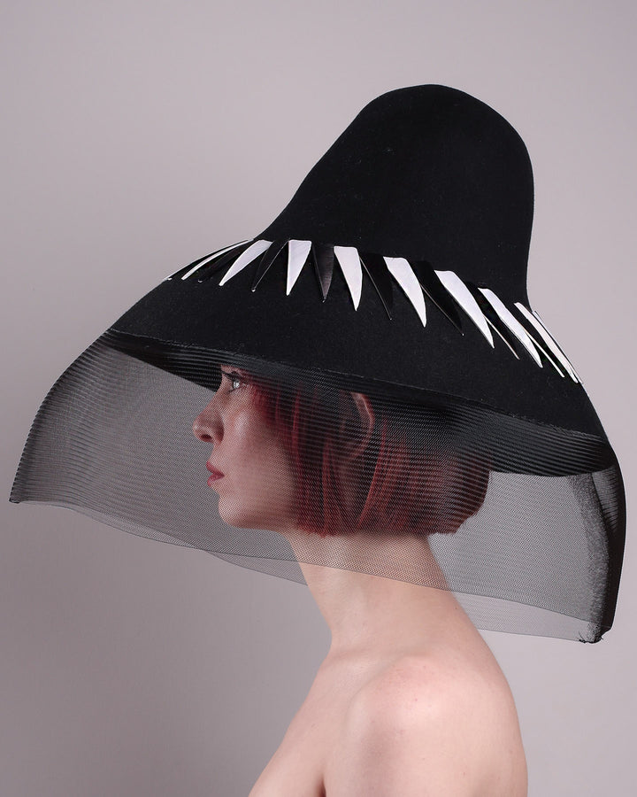 The model showcasing the left side close up of the Ava hat, featuring an exaggerated, proportioned sculptural high-coned silhouette crafted in jet-black felt and crowned with sharp white and black feather accents. A sheer black crinoline veil spills from the brim. Designed by Merve Bayindir.