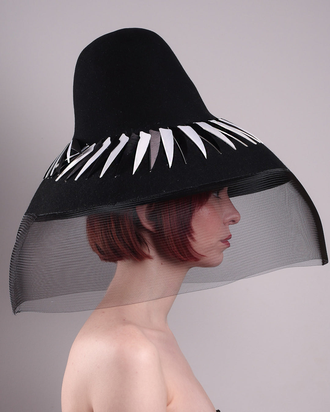 The model showcasing the right side close up of the Ava hat, featuring an exaggerated, proportioned sculptural high-coned silhouette crafted in jet-black felt and crowned with sharp white and black feather accents. A sheer black crinoline veil spills from the brim. Designed by Merve Bayindir.