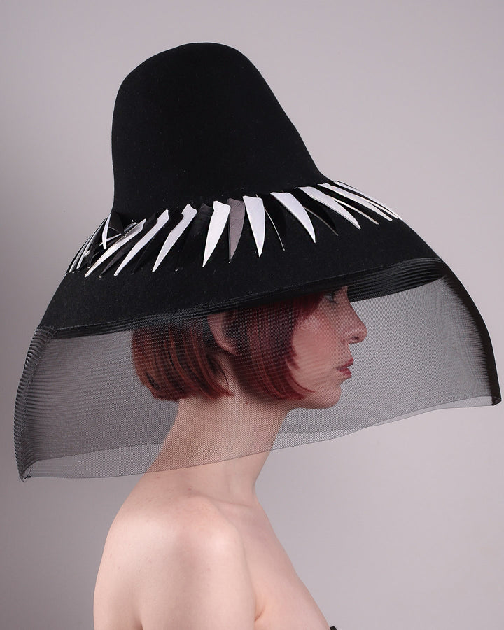 The model showcasing the right side close up of the Ava hat, featuring an exaggerated, proportioned sculptural high-coned silhouette crafted in jet-black felt and crowned with sharp white and black feather accents. A sheer black crinoline veil spills from the brim. Designed by Merve Bayindir.