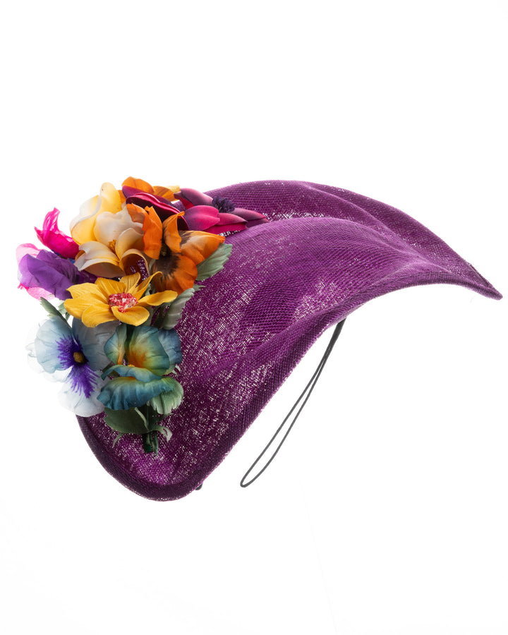 Purple sinamay sculpted fascinator with a curved, wave-like shape, adorned with a vibrant cluster of multicolored flowers and mounted on a slim black headband. Designed and made by Merve Bayindir Millinery.

