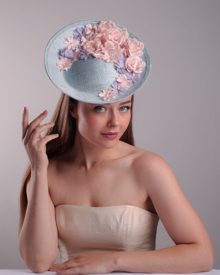 The model is showcasing the front of a blue classic saucer hat with curved hight on the back left worn towards the right side of the head. The top of the hat is decorated with pink and purple flower&amp;nbsp;arrangement. The hat is fitted with a wire headband and an elastic.